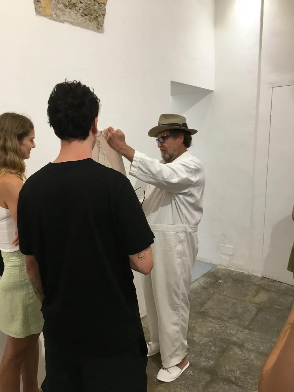Julian Schnabel, in completo bianco e cappello, osserva da vicino un piccolo dipinto tenuto in mano, tra alcuni visitatori
